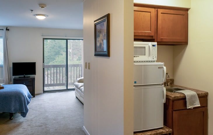 An apartment interior featuring a compact kitchen with wooden cabinets and appliances, opening to a living room with a balcony view.