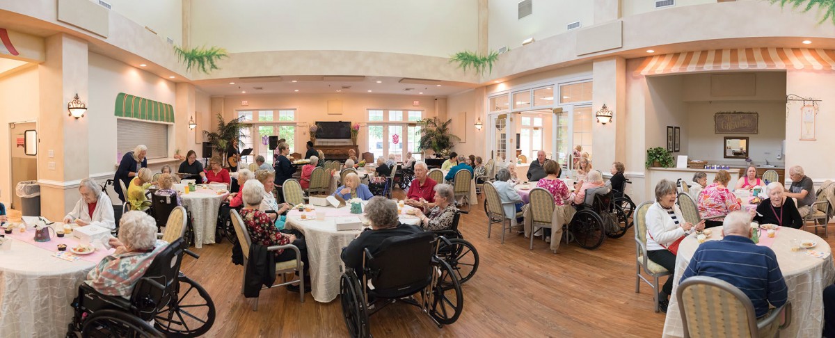 A group of seniors gathered in a dining hall, some using wheelchairs, enjoying a communal event.