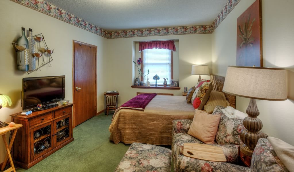A cozy bedroom with a neatly made bed, patterned chair, and decorative elements on the window sill.