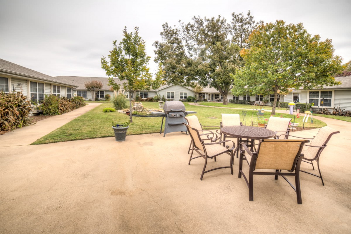A spacious outdoor patio area with a table, chairs, and a grill surrounded by a lush garden.