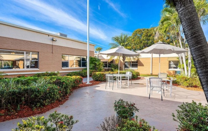 A sunny outdoor patio area with tables, chairs, and umbrellas surrounded by a landscaped garden.