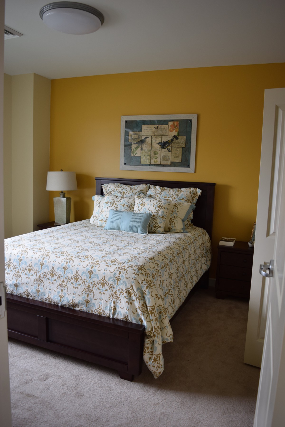 A cozy bedroom featuring a bed with patterned bedding, a bedside lamp, and artwork on a yellow accent wall.