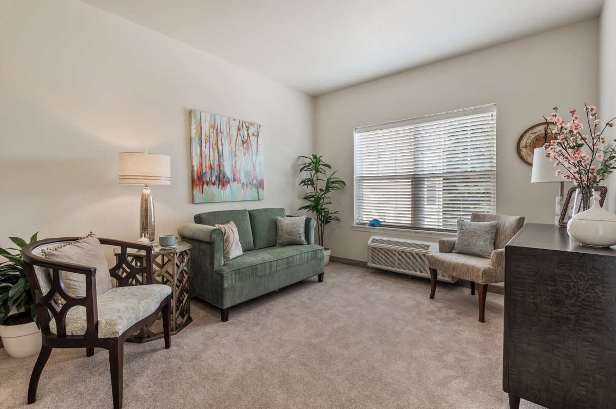 A cozy living room with modern decor, featuring a green sofa, chairs, floor lamp, artwork, and large window.