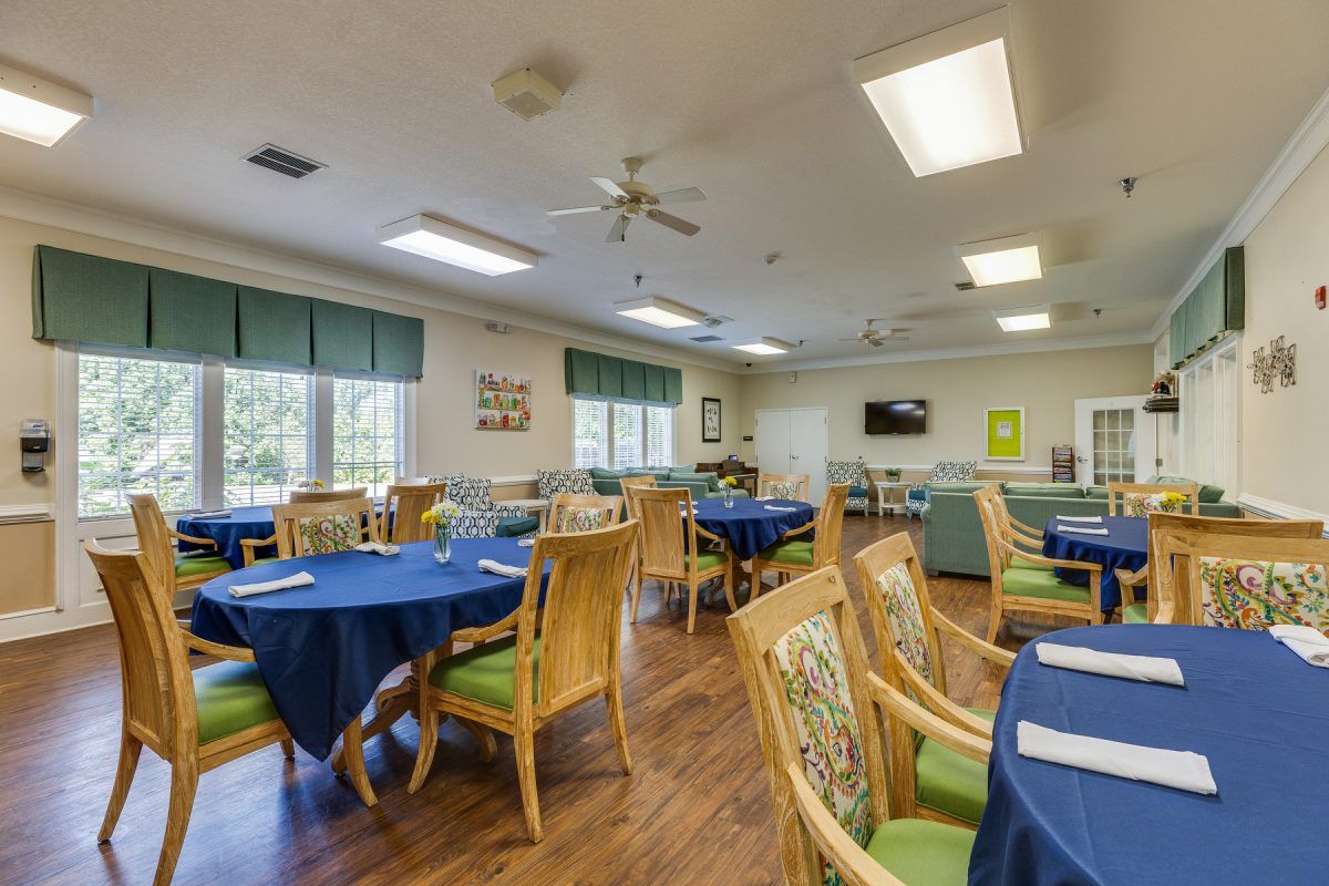 A spacious dining room with blue tablecloths, wooden chairs, and ample natural lighting from large windows.