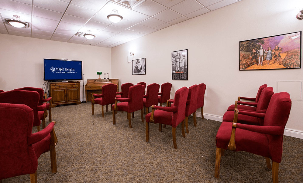A small conference room with red upholstered chairs arranged in rows, a television, and framed pictures on the walls.