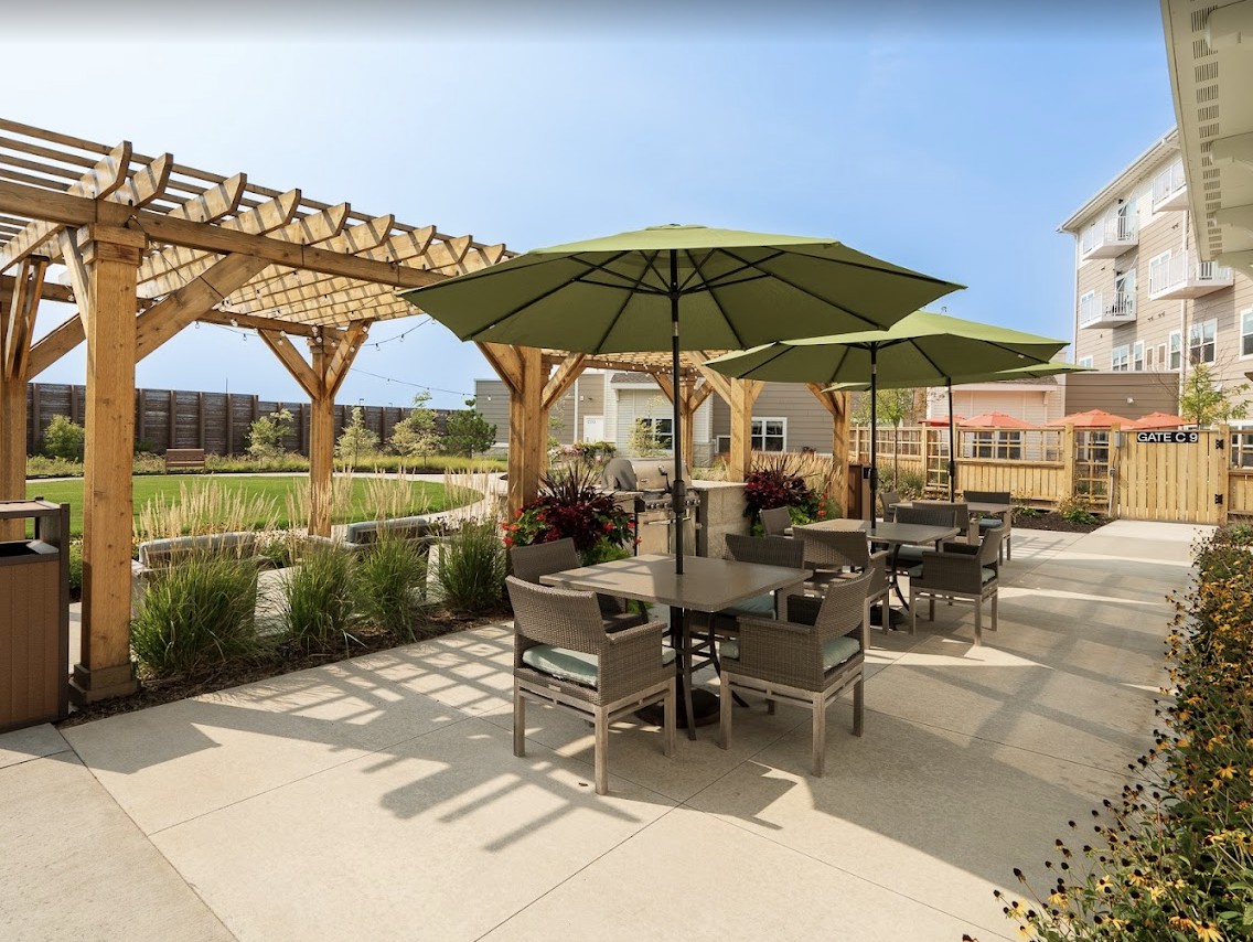 A sunny outdoor patio with tables, chairs, and umbrellas under a wooden pergola.
