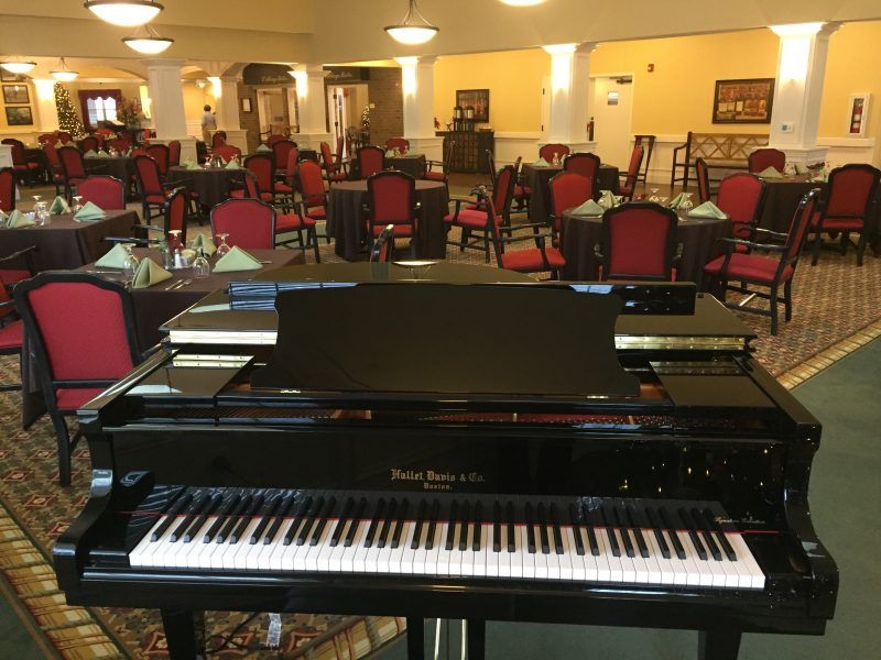 A black piano placed in an empty restaurant with red and black chairs.