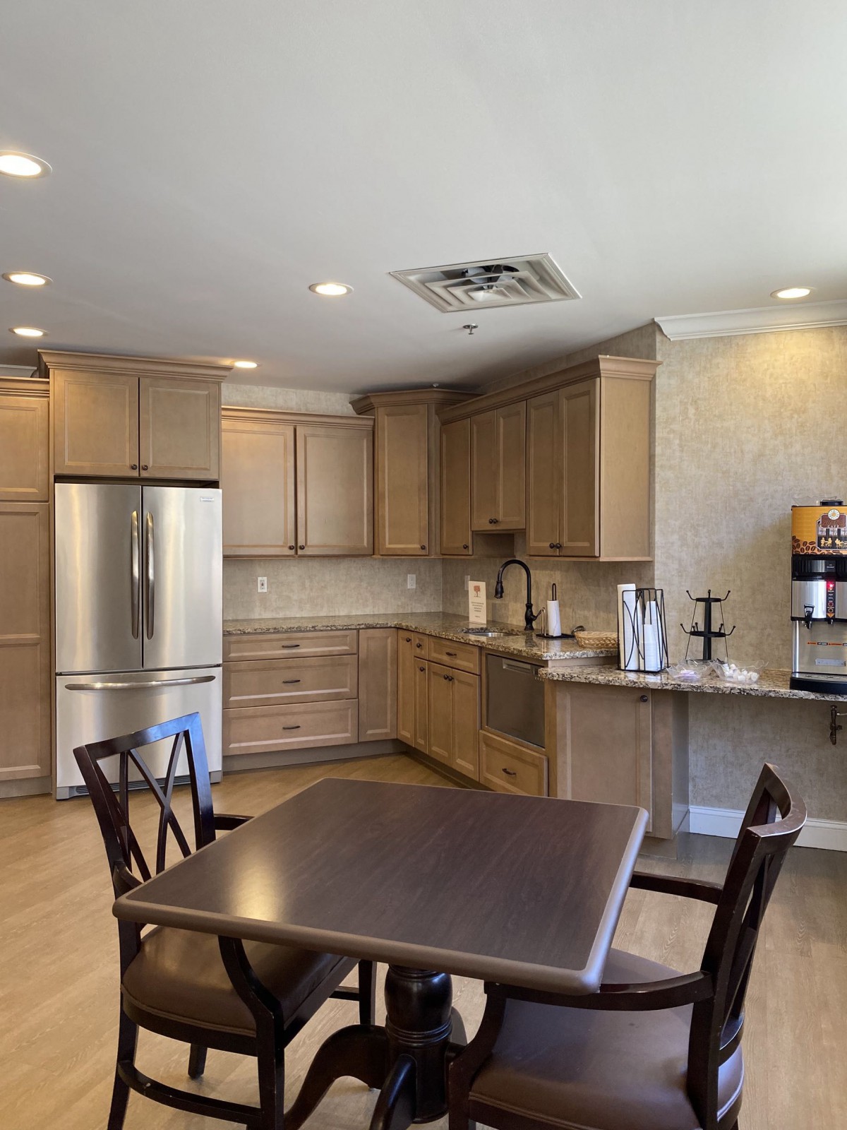 A modern kitchen interior featuring wooden cabinets, a dining table with chairs, a refrigerator, and a coffee machine.
