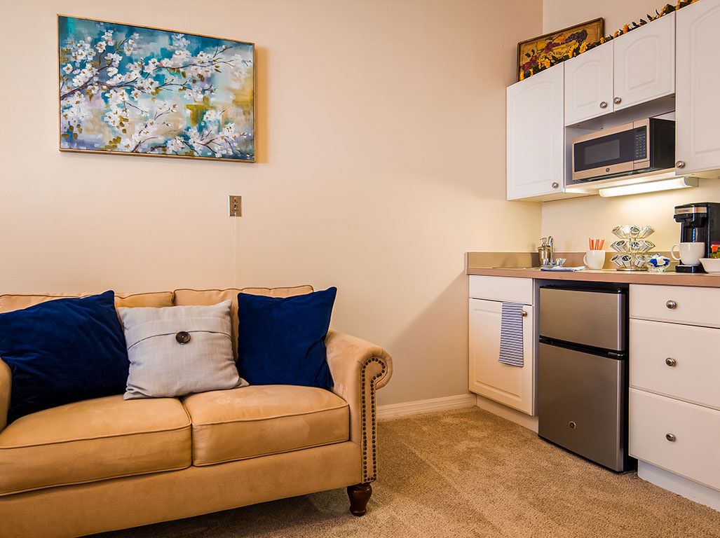 A cozy living area featuring a beige sofa with blue pillows next to a kitchenette, adorned with a piece of abstract artwork.