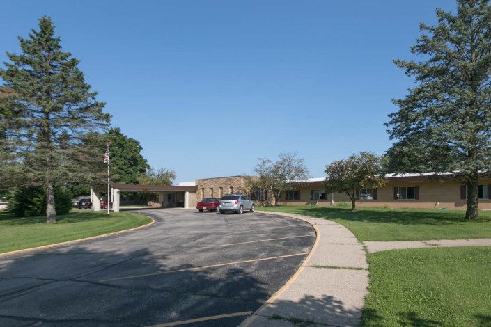 A school building with a curved driveway and parked cars surrounded by trees under a clear blue sky.