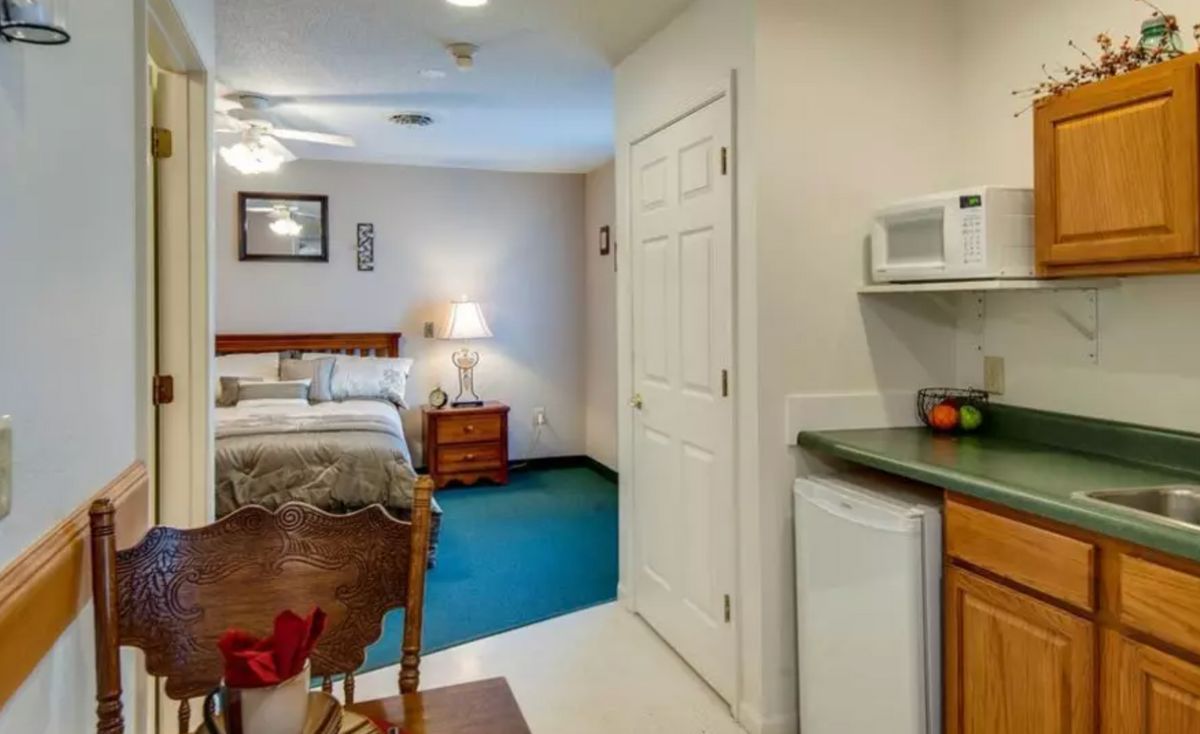 A cozy hotel room featuring a bed, nightstand with a lamp, and a small kitchenette area.
