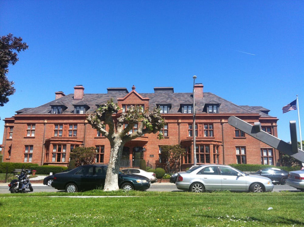 A large brick building with parked cars and a tree in front under a clear blue sky.