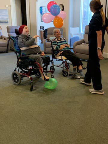 Two elderly individuals in wheelchairs are engaged in an activity with a caregiver in a cheerful indoor setting.