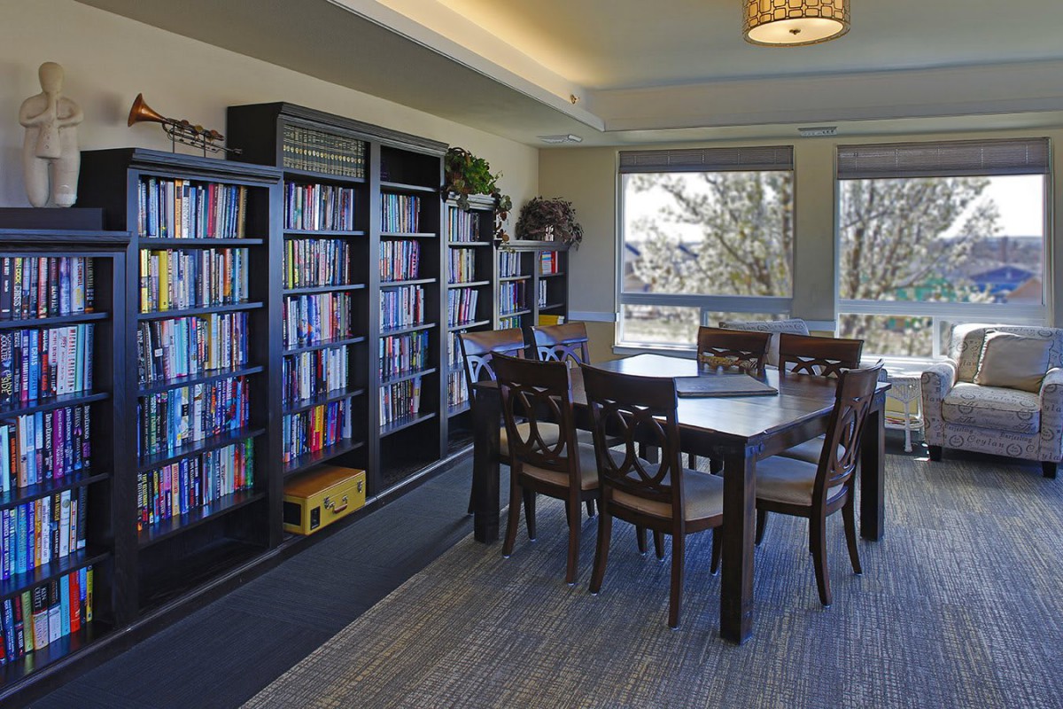 A cozy reading room with bookshelves, a table, and chairs by large windows.