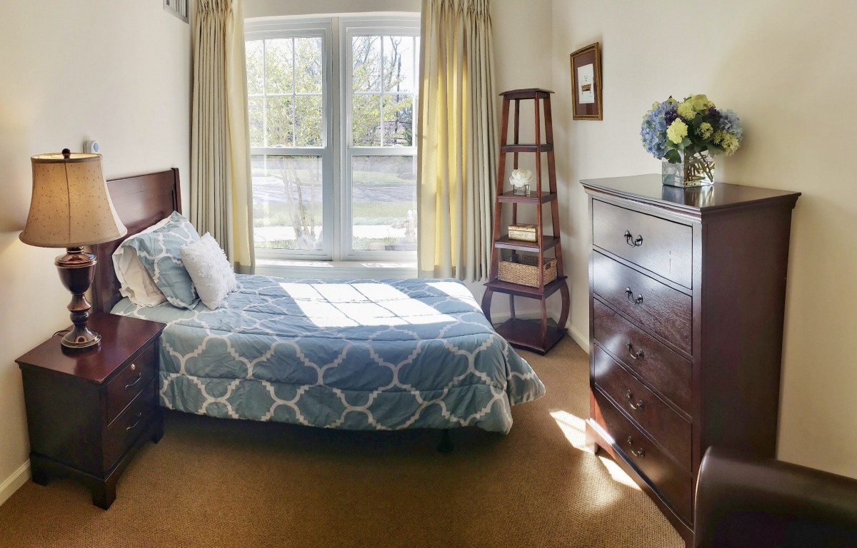 A cozy bedroom with a large window letting in natural light, featuring a bed with blue patterned bedding, a wooden dresser, and a corner shelf.