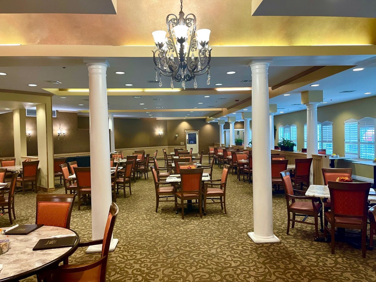 An elegant dining room with arranged tables and chairs, featuring a chandelier and decorative pillars.