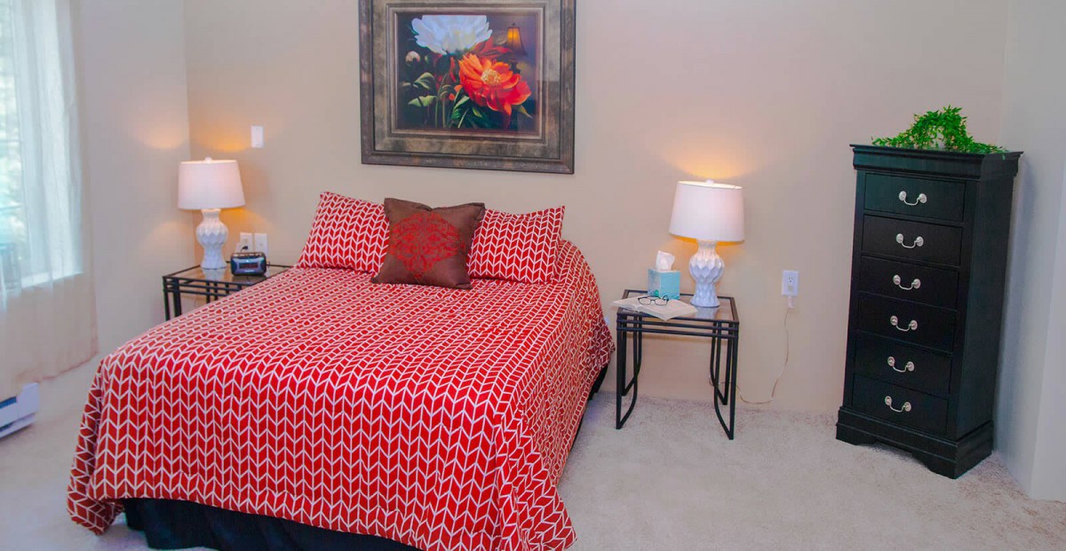 A cozy bedroom with a red patterned bedspread, a stylish nightstand with lamps, and artwork on the wall.