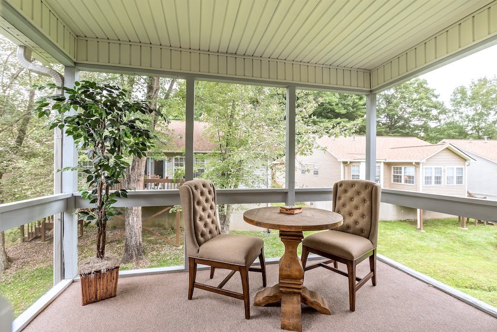 A cozy sunroom with a round table and two upholstered chairs, featuring large windows and indoor plants.