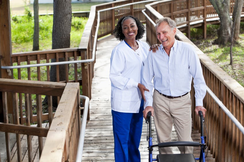 A nurse assists an elderly man with a walker on a wooden pathway outdoors.