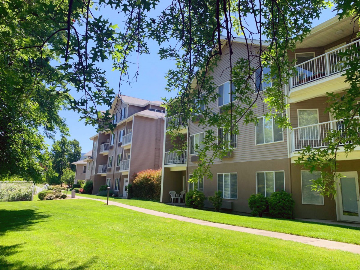 A view of apartment buildings surrounded by green lawns and trees under a clear blue sky.