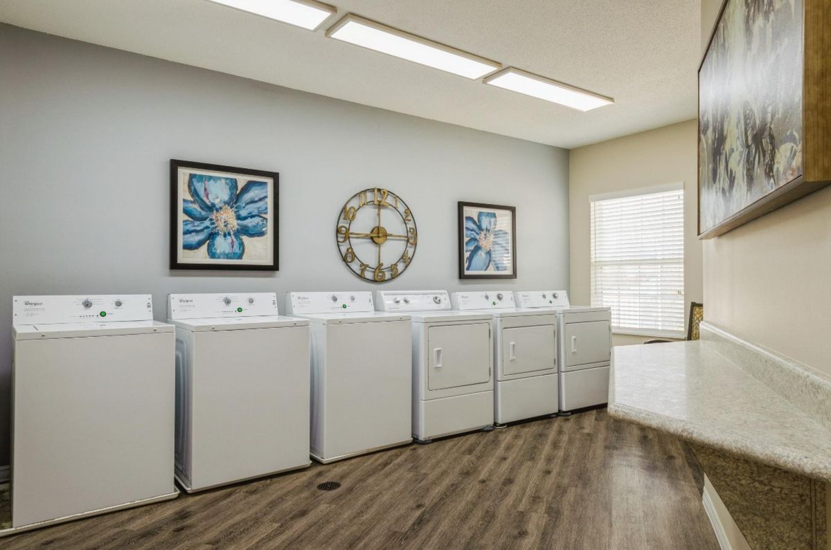 A bright laundry room featuring multiple washing machines and dryers with decorative artwork and a wall clock.