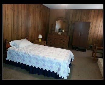 A modest bedroom with wood-paneled walls, featuring a single bed and a dresser with a mirror.