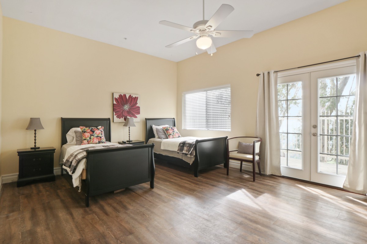 A spacious bedroom with two twin beds, wooden flooring, a ceiling fan, and ample natural light from a large window and door.