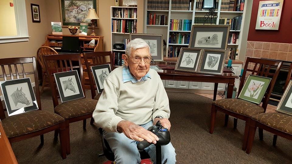 An elderly man sits in a library with framed art pieces on display behind him.
