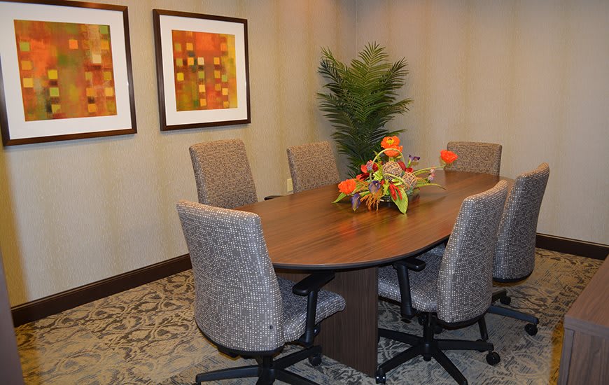 A conference room with a wooden meeting table, surrounded by upholstered chairs, artwork on the walls, and a decorative plant.