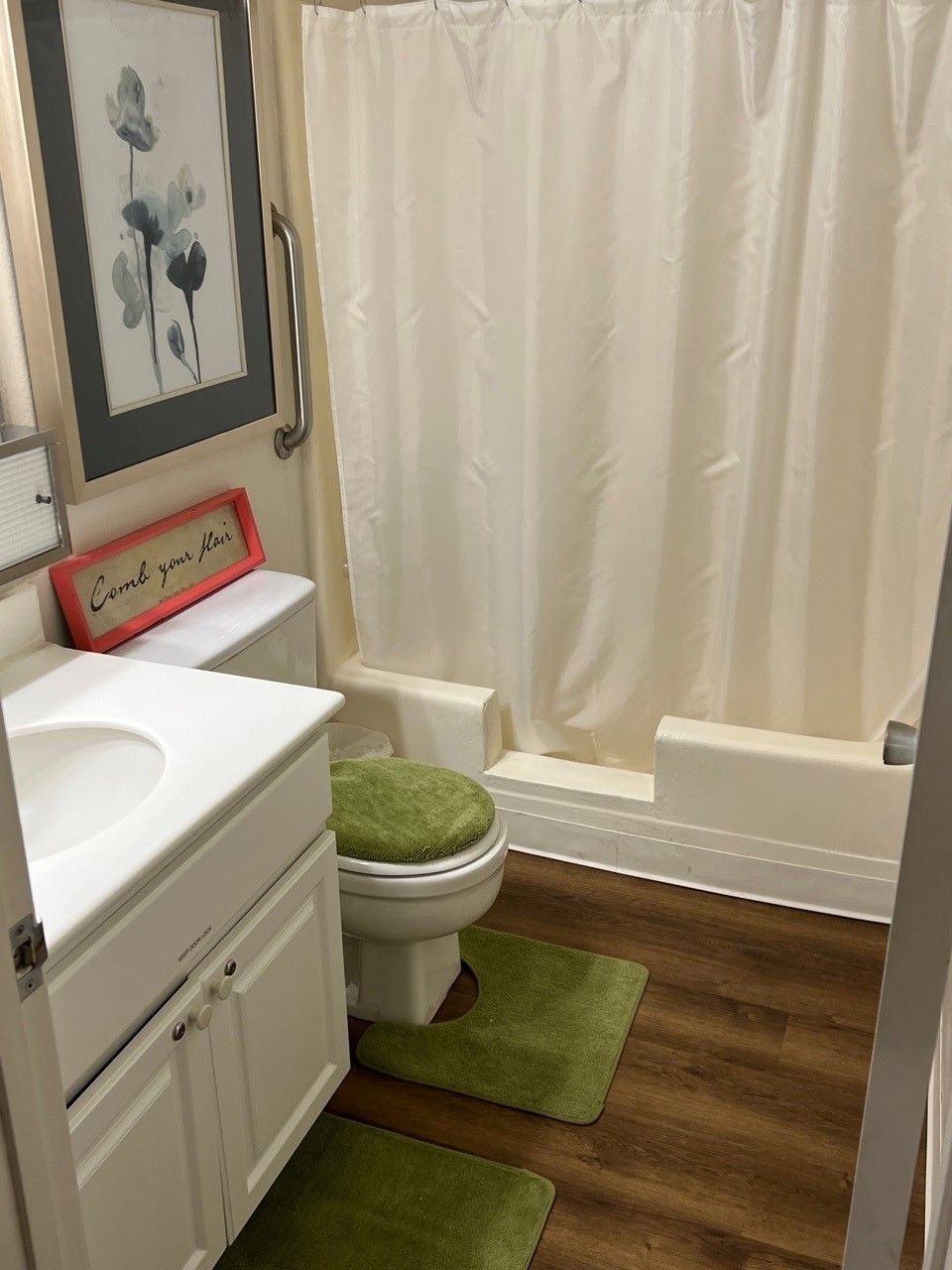 A cozy bathroom with a white sink, toilet, and shower curtain, featuring green mats and wooden flooring.