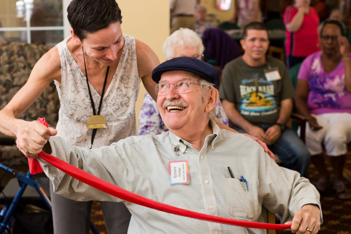 An elderly man joyfully participates in a ribbon-cutting ceremony with others in a community setting.