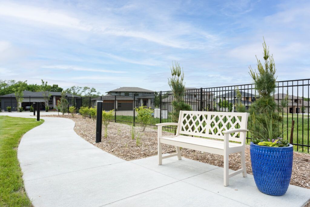 A white bench placed alongside a garden pathway with surrounding greenery and a black metal fence.
