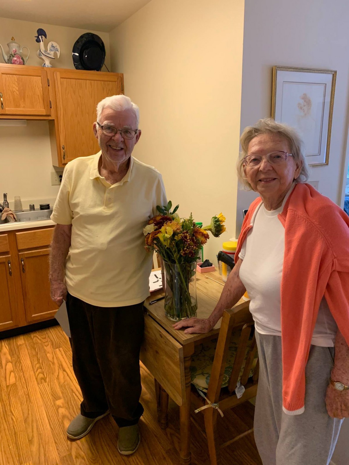 An elderly couple smiling in their home near a bouquet of flowers.