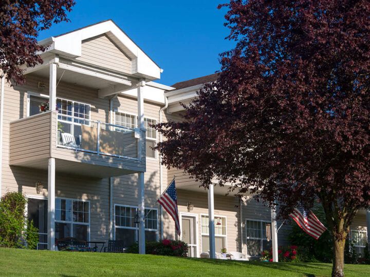A residential apartment building with balconies, surrounded by trees and featuring American flags against a clear blue sky.