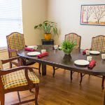 A cozy dining room featuring a wooden table with chairs, adorned with plates and red napkins, complete with a potted plant centerpiece and wall artwork.