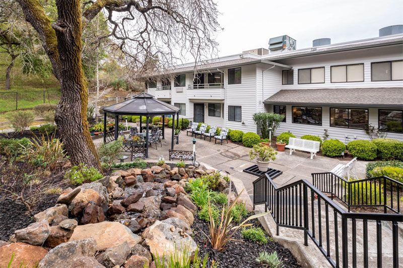 A serene outdoor garden with a gazebo and seating area, surrounded by lush greenery and a rock feature in front of a two-story building.