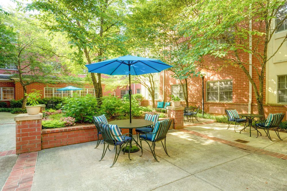 A peaceful outdoor patio with a table, chairs, and blue umbrella surrounded by greenery and a brick building.