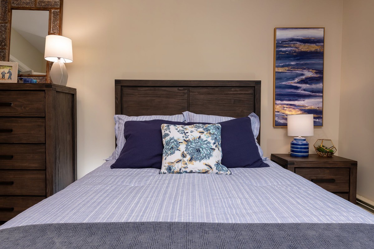 A neatly made bed with decorative pillows in a cozy bedroom, featuring a nightstand and a wall painting.