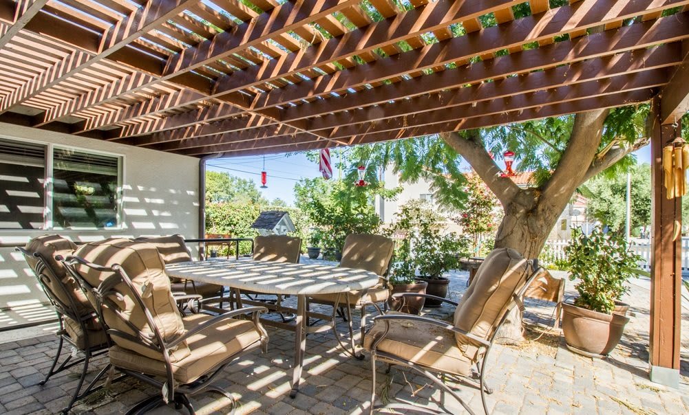 A cozy outdoor dining area under a wooden pergola with comfortable seating and surrounded by greenery.