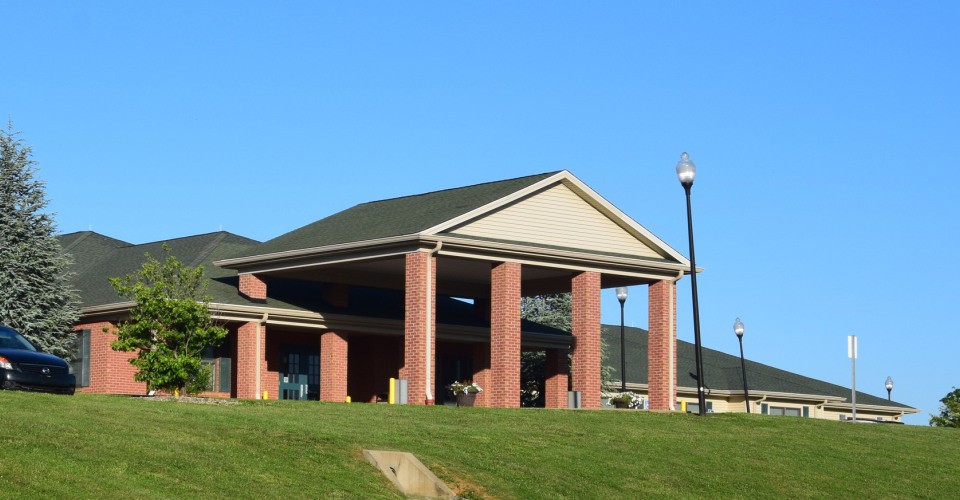 A brick building with columns and a sloped roof, set against a clear blue sky and surrounded by a green lawn.