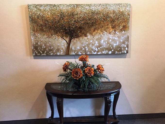 A decorative table with a floral arrangement sits beneath a painting of a tree.