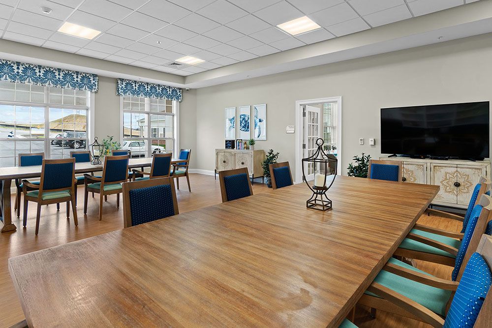 A modern conference room with a large wooden table and multiple chairs, featuring a television and windows allowing natural light.