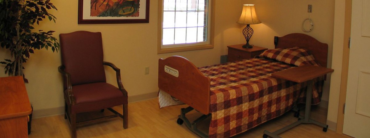 A well-lit room with a hospital bed covered in a checkered quilt, alongside a chair and a lamp.