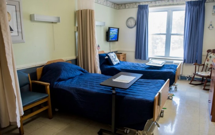 A hospital room with two patient beds, a curtain for privacy, a television on the wall, and a window providing natural light.