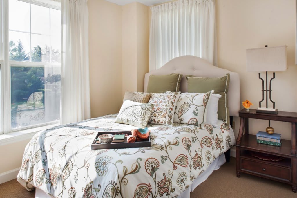 A cozy bedroom with a neatly made bed featuring floral-patterned bedding, a headboard, and decorative pillows, situated near a window with sheer curtains. A tray with items is placed on the bed.