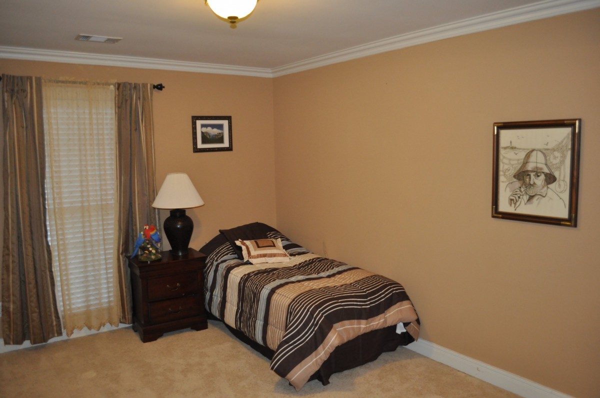 A cozy bedroom with a single bed and a nightstand featuring a lamp, set against beige walls and a carpeted floor.