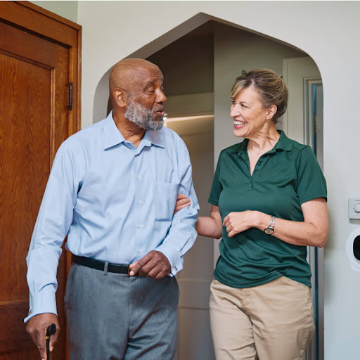 A caregiver assists an elderly man with a friendly conversation as they walk indoors.