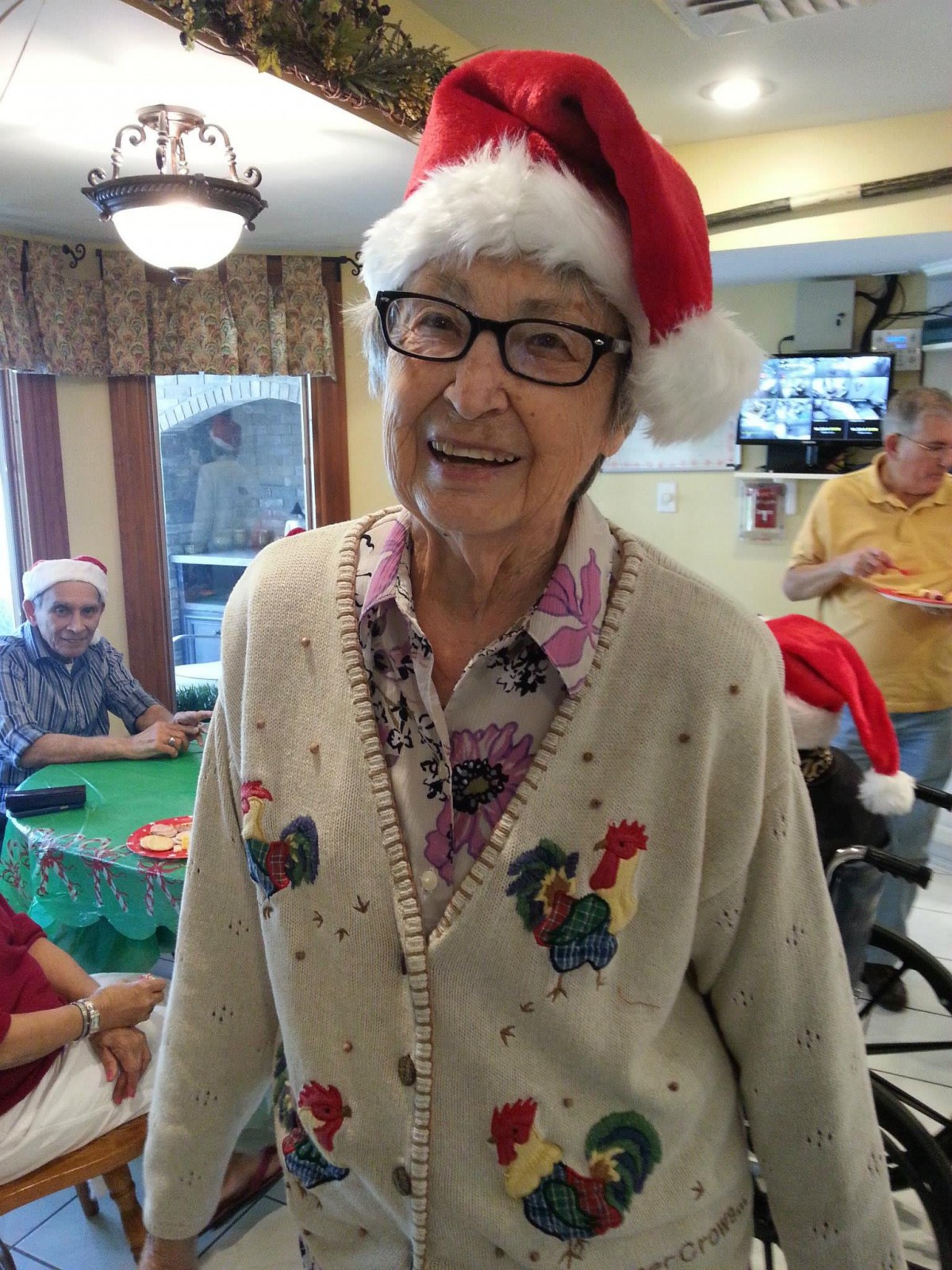 An elderly person wearing a festive Santa hat and sweater smiles indoors.