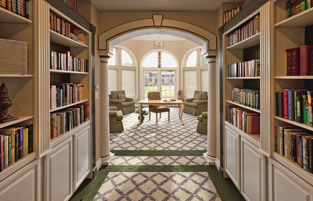 A cozy reading area with bookshelves, comfortable chairs, and a table, illuminated by natural light through large windows.