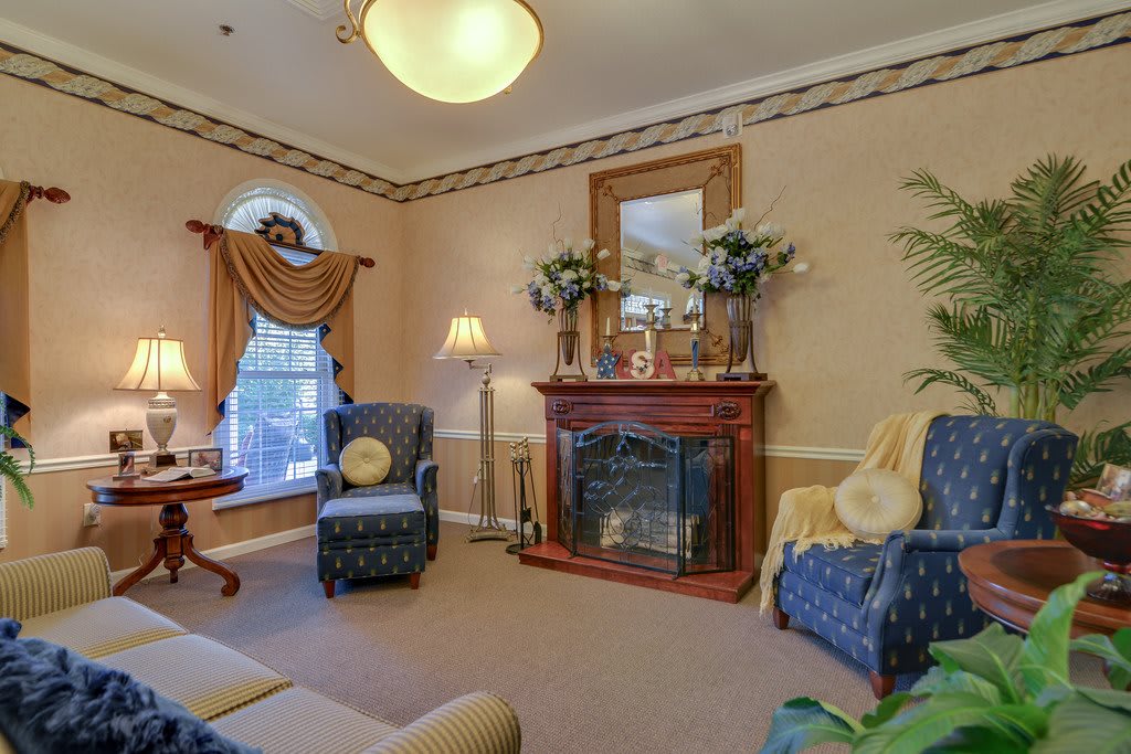 A traditional living room featuring a fireplace, two blue armchairs, elegant drapes, and decorative elements such as a mirror and plants.